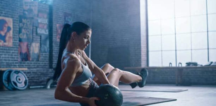 Close Up of a Strong and Fit Athletic Woman in Sport Top Doing Core and Ab Exercises with Ball in a Loft Style Industrial Gym with Motivational Posters. It's Part of Her Cross Fitness Training Workout